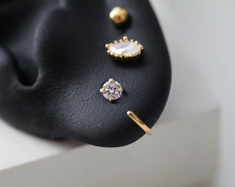 May include: Close-up of a black ear with multiple earrings. The earrings include a small gold ball, a teardrop-shaped earring with a gold frame and a clear stone, a round clear stone stud, and a gold hoop earring.