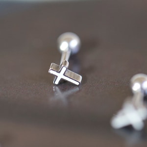 May include: Silver-colored cross earrings with a ball backing. The cross design is detailed and the earrings are small. The earrings are displayed on a dark surface.