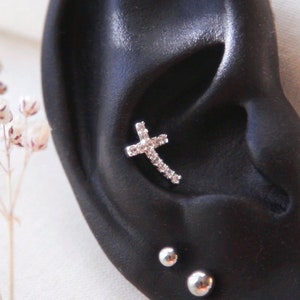 May include: A black ear model with a silver cross earring with clear cubic zirconia stones. The ear model also has two silver ball earrings.