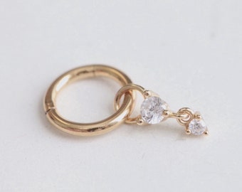 May include: A gold-colored hinged hoop earring with a dangling charm. The charm features two clear, sparkling, teardrop-shaped cubic zirconia stones. The earring is set against a white background.