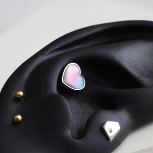 Puede incluir: Primer plano de un modelo de oreja negro con tres pendientes diferentes. El pendiente del centro es una gema en forma de corazón con un acabado iridiscente rosa, azul y blanco. Los otros dos pendientes son pequeñas bolas doradas.