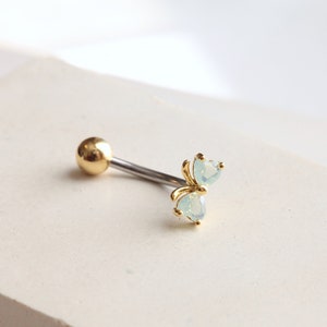 May include: A gold-colored barbell-style body piercing with a small, round ball on one end and a decorative cluster of two heart-shaped, light blue gemstones on the other. The jewelry is set against a white background.