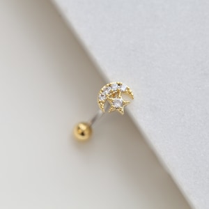 May include: Gold-toned earring featuring a crescent moon and star design, embellished with clear gemstones. The earring has a small gold ball at the end of the post. The jewelry is set against a white background.