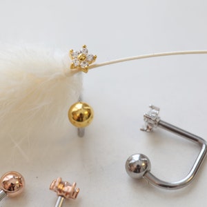 May include: A collection of body jewelry, including a gold-colored flower-shaped stud earring with clear stones, a gold ball stud, and several curved barbell earrings with ball and flower-shaped ends. The jewelry is displayed on a white surface.