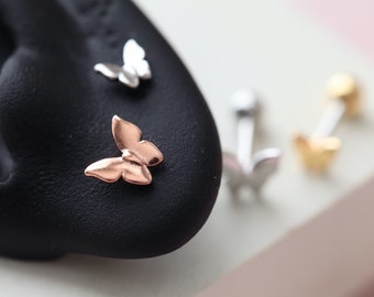 Könnte beinhalten: Nahaufnahme mehrerer ohrringförmiger Schmetterlinge. Einer ist roségoldfarben, ein anderer silberfarben. Die Ohrringe befinden sich auf einer schwarzen Oberfläche. Die Schmetterlingsohrringe sind ein zartes und stilvolles Accessoire.