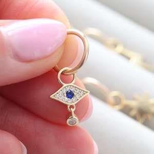 May include: Gold-colored hoop earring with an eye-shaped charm. The charm features a blue center stone surrounded by smaller white stones. A small, dangling clear stone hangs below the eye. Other gold earrings are visible in the background.