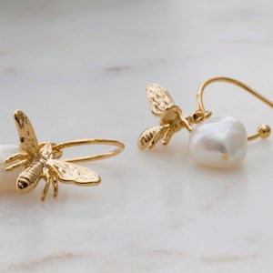 Pode incluir: Brincos de abelha dourados com gotas de pérolas brancas. Os brincos apresentam pingentes de abelha detalhados presos a um aro e uma pérola. O design é elegante e adequado para uso diário ou ocasiões especiais.