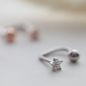 Puede incluir: Un aro de nariz plateado con una pequeña piedra preciosa en forma de flor transparente. El aro es curvo y tiene una pequeña bola en el extremo.