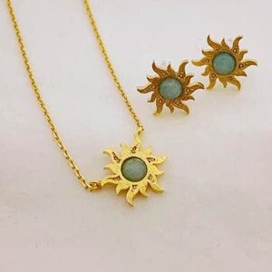 May include: A gold-colored necklace and matching earrings set. The pendant and earrings feature a sunburst design with a central, light-blue stone. The jewelry is displayed on a white surface.