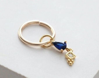 May include: A gold-colored hoop earring with a dangling charm. The charm features a teardrop-shaped, dark blue gemstone and a small square with a clear stone. The earring is set against a white background.