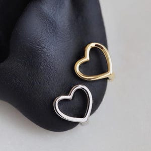 May include: Two heart-shaped ear cuffs, one gold-toned and one silver-toned, are displayed on a black surface. The open-heart design is a simple, yet elegant jewelry piece. The ear cuffs are a modern take on ear jewelry.