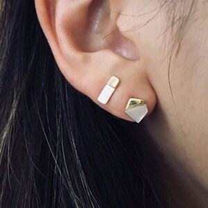 May include: Close-up of a person's ear with two gold earrings. One earring is a rectangular shape with a white enamel inlay. The other earring is a geometric shape with a white enamel inlay. The earrings are set against dark hair.