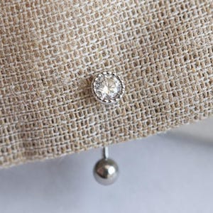 Può includere: Primo piano di un tessuto beige con un bottone decorativo rotondo argentato con una pietra centrale chiara e scintillante. Una piccola sfera argentata è appesa sotto.
