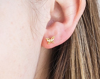 Könnte beinhalten: Nahaufnahme eines Ohrs mit einem kleinen, goldenen Schmetterlings-Ohrstecker. Der Schmetterling hat detaillierte Flügel und einen kleinen, klaren Stein. Der Ohrring ist vor der Haut und den Haaren der Person platziert.