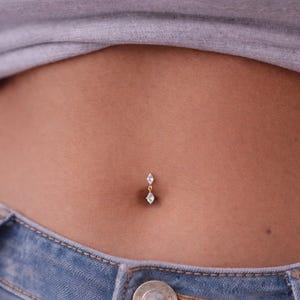 Könnte beinhalten: Ein goldfarbener Bauchnabelpiercing mit zwei klaren, marquiseförmigen Edelsteinen. Der Schmuck hängt an einer kleinen Kette und ist vor Haut und blauen Jeans abgebildet.