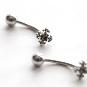 May include: Two curved, silver-colored body jewelry pieces. Each piece has a small, round ball on one end and a decorative, fleur-de-lis shaped charm with a dark center stone on the other end. The jewelry is set against a white background.