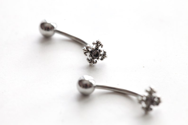 May include: Two curved, silver-colored body jewelry pieces. Each piece has a small, round ball on one end and a decorative, fleur-de-lis shaped charm with a dark center stone on the other end. The jewelry is set against a white background.