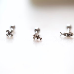 May include: Three silver-toned earrings with decorative tops. The designs include a snowflake, a fleur-de-lis, and a fish. Each earring has a small, round post. The earrings are displayed on a white surface.