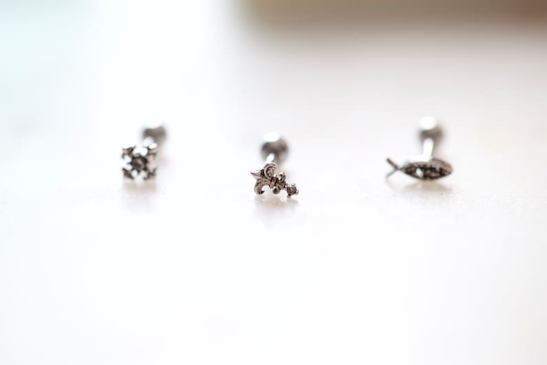 May include: Three silver-toned earrings with decorative tops. The designs include a snowflake, a fleur-de-lis, and a fish. Each earring has a small, round post. The earrings are displayed on a white surface.