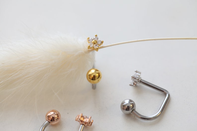 May include: A collection of body jewelry, including a gold-colored flower-shaped stud earring with clear stones, a gold ball stud, and several curved barbell earrings with ball and flower-shaped ends. The jewelry is displayed on a white surface.
