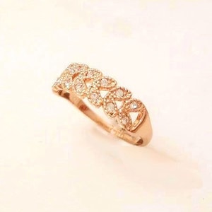 May include: A rose gold ring with a repeating pattern of openwork shapes, each set with small, clear gemstones. The ring is displayed on a light-colored surface, showcasing its intricate design and sparkle.
