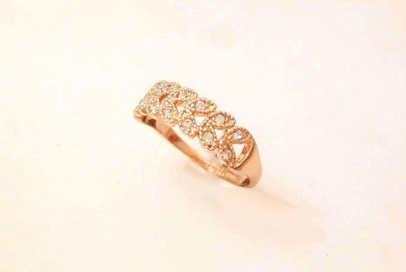 May include: A rose gold ring with a repeating pattern of openwork shapes, each set with small, clear gemstones. The ring is displayed on a light-colored surface, showcasing its intricate design and sparkle.