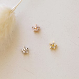 May include: Three small, star-shaped earrings with a floral design. One is silver, one is rose gold, and one is gold. Each earring features small, clear gemstones. The earrings are displayed on a white surface.