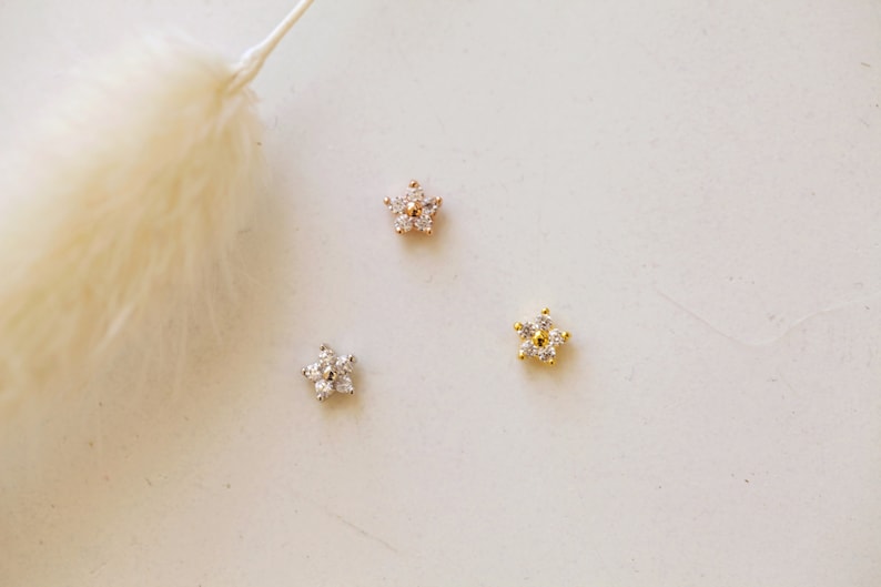 May include: Three small, star-shaped earrings with a floral design. One is silver, one is rose gold, and one is gold. Each earring features small, clear gemstones. The earrings are displayed on a white surface.
