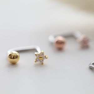 May include: Close-up of several pieces of jewelry. One piece is a silver-colored curved barbell with a gold-colored ball on one end and a gold-colored flower with clear stones on the other. Other pieces are visible in the background.