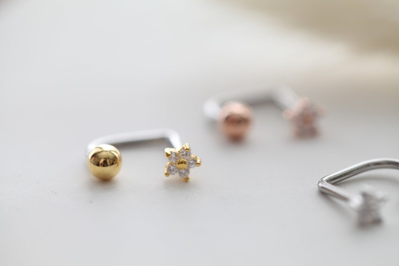 May include: Close-up of several pieces of jewelry. One piece is a silver-colored curved barbell with a gold-colored ball on one end and a gold-colored flower with clear stones on the other. Other pieces are visible in the background.