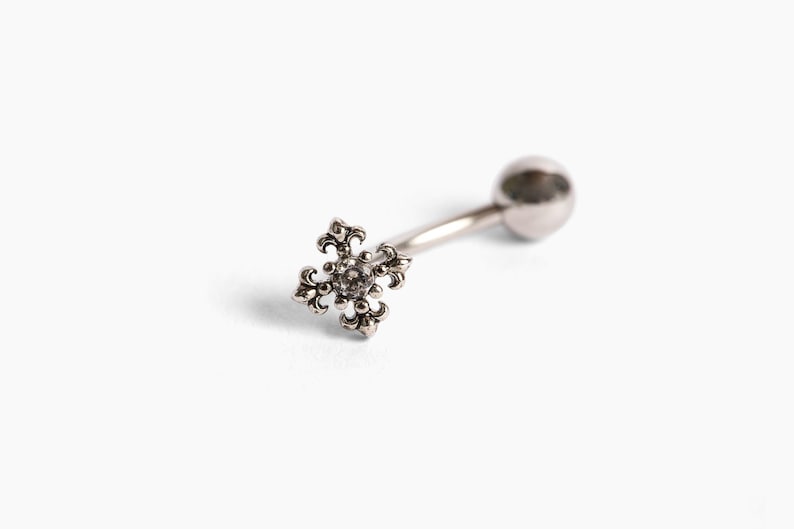 May include: A silver-colored belly button ring with a decorative cross-shaped charm. The charm features an ornate, fleur-de-lis design with a small, clear gem in the center. The barbell is curved with a ball on one end.