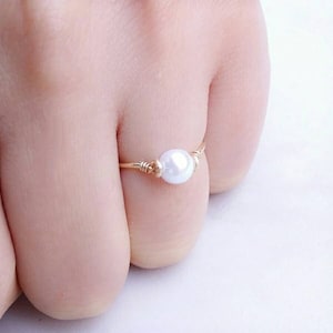 May include: A delicate gold-toned ring featuring a single, luminous white pearl. The pearl is centered on the ring, held in place by intricate wire wrapping. The ring's band is thin and elegant, showcasing the pearl's natural beauty.