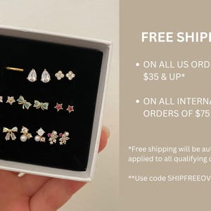 May include: A white jewelry box with a black velvet insert containing twelve gold-tone stud earrings. The collection includes teardrop, flower, bow, and star shapes, accented with clear crystals, small white pearls, and pink stones.