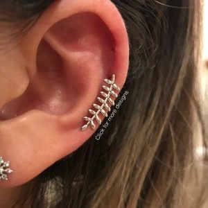Puede incluir: Un pendiente de oreja plateado con forma de rama de hojas, usado en la oreja. El pendiente tiene una serie de hojas pequeñas y detalladas. El texto "Click for more designs" es visible en la imagen.