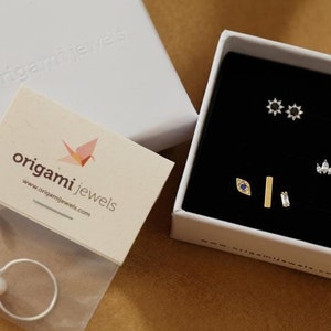 May include: A jewelry set with various earrings and a ring. The earrings are in a black box, including starburst, teardrop, and bar designs. A ring with a pearl is in a plastic bag. The brand name "origami jewels" is visible.
