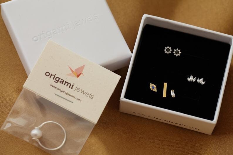 May include: A jewelry set with various earrings and a ring. The earrings are in a black box, including starburst, teardrop, and bar designs. A ring with a pearl is in a plastic bag. The brand name "origami jewels" is visible.