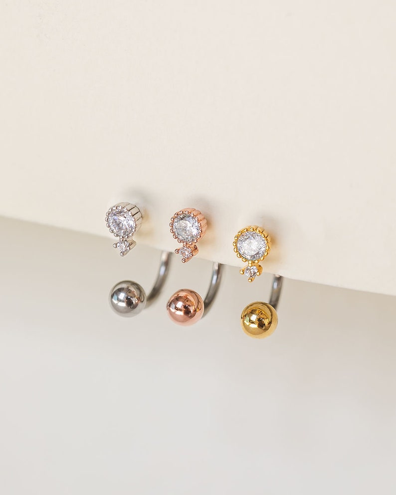 Pu&ograve; includere: Tre orecchini a doppia faccia in argento, oro rosa e oro. Ogni orecchino presenta una piccola gemma rotonda e trasparente nella parte superiore, con una gemma pi&ugrave; piccola sottostante. La parte inferiore di ogni orecchino ha una sfera di metallo abbinata.