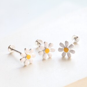 May include: Three stud earrings featuring a daisy flower design with white petals and a yellow center. Each earring has a silver-toned post and a flat circular backing, showcasing a delicate floral motif with a clean, polished finish.