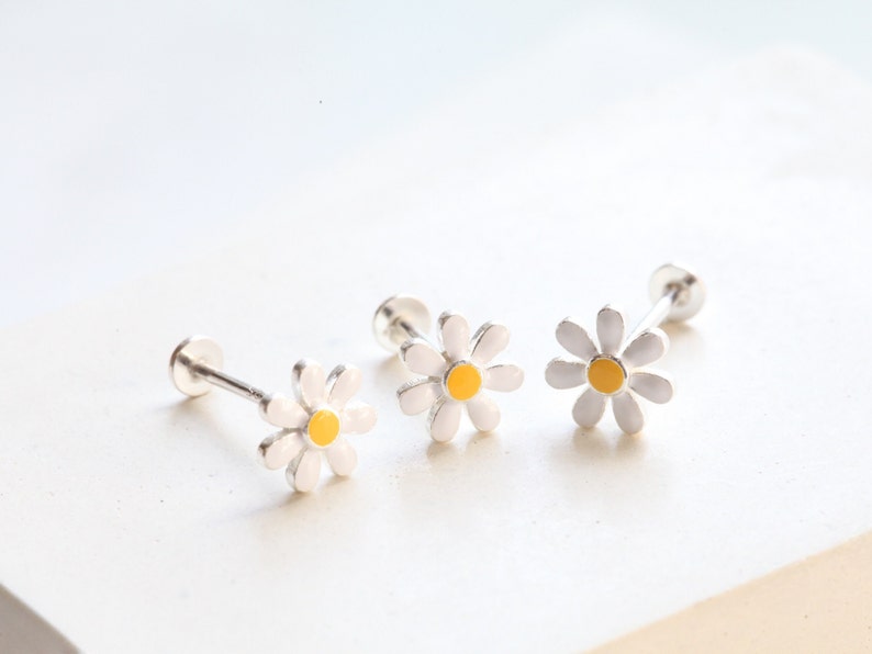 May include: Three stud earrings featuring a daisy flower design with white petals and a yellow center. Each earring has a silver-toned post and a flat circular backing, showcasing a delicate floral motif with a clean, polished finish.
