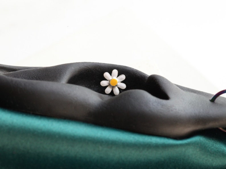 May include: A small daisy-shaped stud earring featuring white petals and a bright yellow center. The earring is positioned on a dark, matte, ear-shaped display stand that rests against a smooth, teal-colored fabric base.