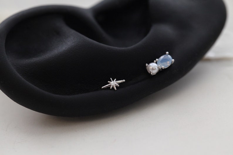 May include: Three silver earrings on a black surface. One is a starburst design, and the other two are a pearl and a light blue teardrop-shaped stone. The earrings are small and delicate.