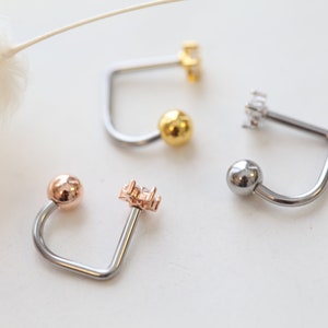 May include: Close-up of four curved barbell earrings. The earrings feature a ball on one end and a decorative element on the other. Colors include silver, gold, and rose gold. The decorative elements include a cluster of small crystals.