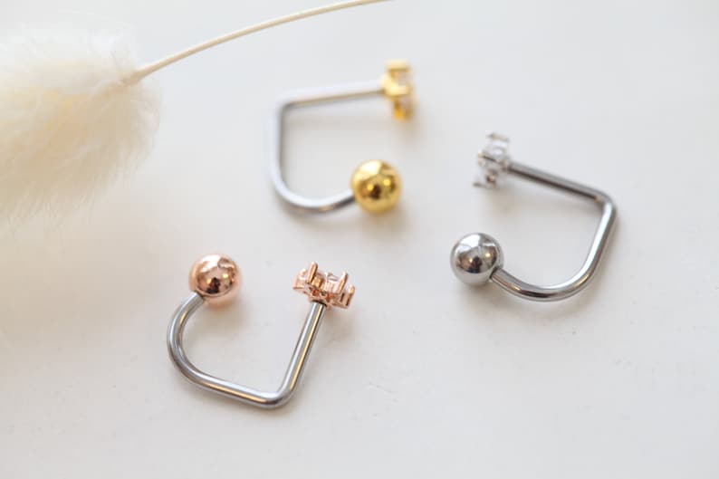 May include: Close-up of four curved barbell earrings. The earrings feature a ball on one end and a decorative element on the other. Colors include silver, gold, and rose gold. The decorative elements include a cluster of small crystals.