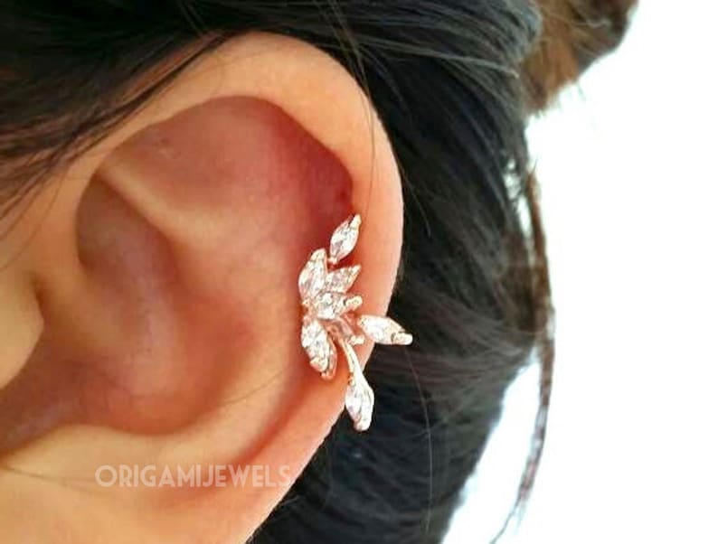 May include: A rose gold ear cuff featuring a leaf design with clear, sparkling crystals. The cuff is worn on the ear and has a delicate, elegant appearance. The brand name "ORIGAMIJEWELS" is visible.