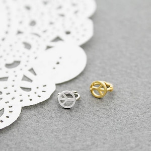 May include: Two small ear cuffs, one silver and one gold, each featuring a peace symbol design. The ear cuffs are displayed on a gray surface with a white, lacy paper doily in the background. The gold ear cuff has a shiny finish.
