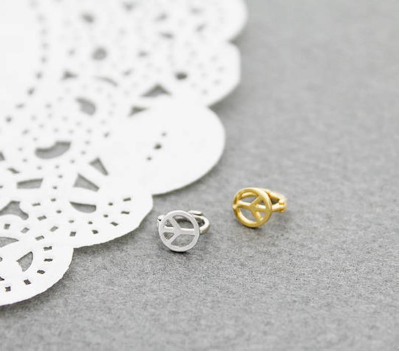 May include: Two small ear cuffs, one silver and one gold, each featuring a peace symbol design. The ear cuffs are displayed on a gray surface with a white, lacy paper doily in the background. The gold ear cuff has a shiny finish.