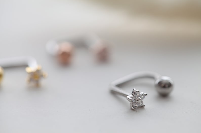 May include: A close-up of a silver horseshoe earring with a small, five-petal flower design encrusted with clear gemstones. The earring has a curved bar and a small ball on the other end.