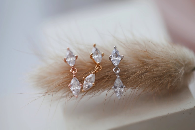 May include: Three pairs of earrings with a dangling marquise-cut cubic zirconia. The earrings are in rose gold, gold, and silver tones. The earrings are displayed on a soft, neutral background.