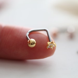 May include: A silver and gold-toned curved barbell earring. One end features a small gold ball, and the other end has a gold star-shaped design with clear, small gemstones. The jewelry is designed for body piercing.