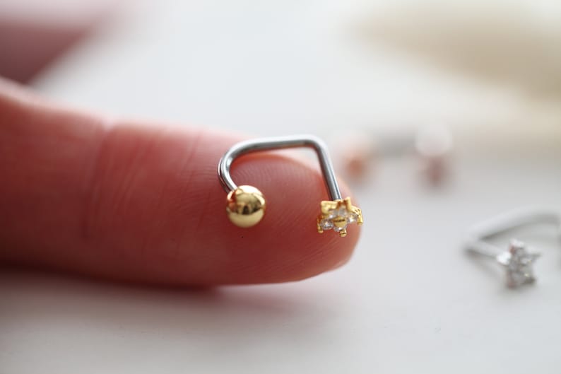 May include: A silver and gold-toned curved barbell earring. One end features a small gold ball, and the other end has a gold star-shaped design with clear, small gemstones. The jewelry is designed for body piercing.